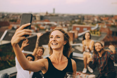 Smiling young woman taking selfie with friends through smart phone on terrace