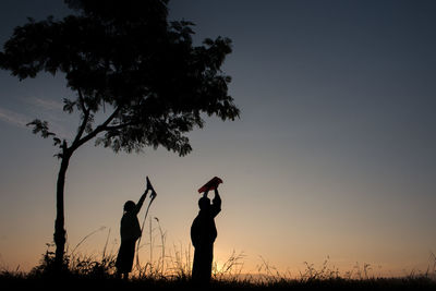 Silhouette children with arms raised against sky during sunset