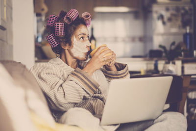 Mature woman with beauty products drinking coffee while using laptop at home