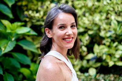 Side view of joyful female with charming toothy smile and brown hair looking at camera on background of green leaves in summer garden