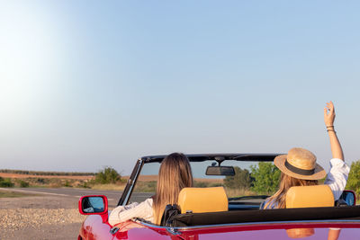 Back view of two friends having fun driving a vintage red car in the countryside during spring break