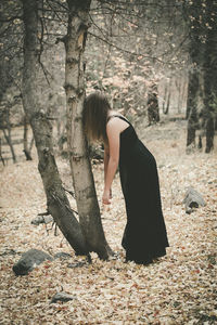 Side view of woman in black dress leaning on bare tree