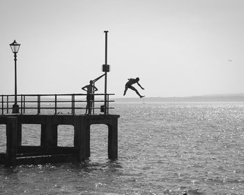 Man standing on pier while looking at friend jumping in sea against clear sky