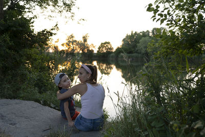 Rear view of friends sitting by lake against trees