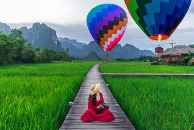 Asian woman holding a camera sits on a wooden walkway and looks at balloons with green rice fields 