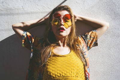 Woman wearing red and yellow sunglasses leaning with hands behind head on wall