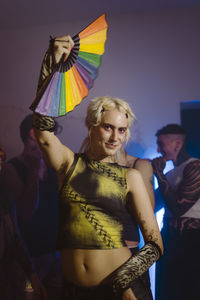 Portrait of smiling non-binary person holding hand fan while standing at nightclub