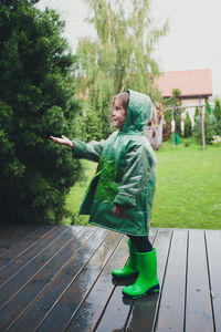 Full length of cute girl wearing raincoat and rubber boots while standing on wet floorboard during rainy season