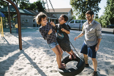 Father looking at children playing on tire swing in backyard