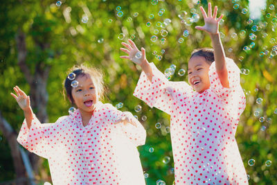 Happy sisters wearing raincoats playing with bubbles against trees in park