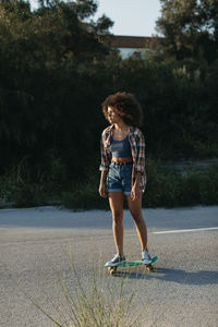 Side view of female hipster with afro hairstyle riding penny board along empty road during sunset