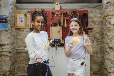 Portrait of smiling female students showing achievements and electrical parts while standing in technology workshop at s