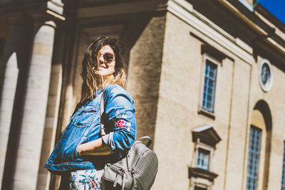 Low angle view of young woman wearing sunglasses with backpack standing against building