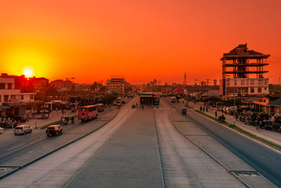 Cars on road by buildings against sky during sunset