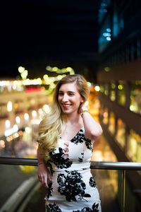 Smiling young woman standing in balcony at illuminated city