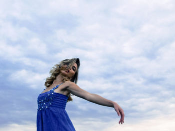 Low angle portrait of beautiful young woman dancing against cloudy sky