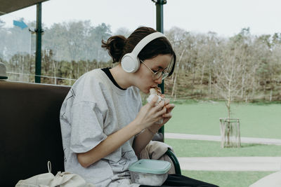 Portrait of one beautiful caucasian teenage girl in white headphones eating, biting a sandwich,