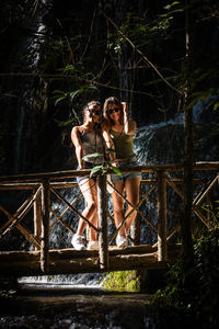 Portrait of smiling friends standing on footbridge over stream in forest