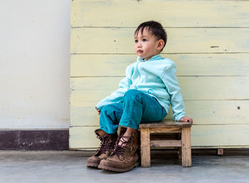 Boy looking away while sitting on seat against wall