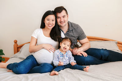 Family asian chinese pregnant woman and caucasian man with toddler girl sitting on bed at home. 