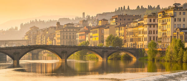 Arch bridge over river by buildings against sky during sunset