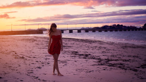 Woman looking down while standing on beach against sky during sunset