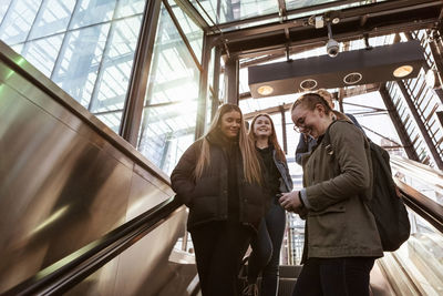 Smiling teenage girl using mobile phone while standing with friends on escalator