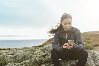 Ireland, howth, female athlete sitting at cliff coast looking at cell phone