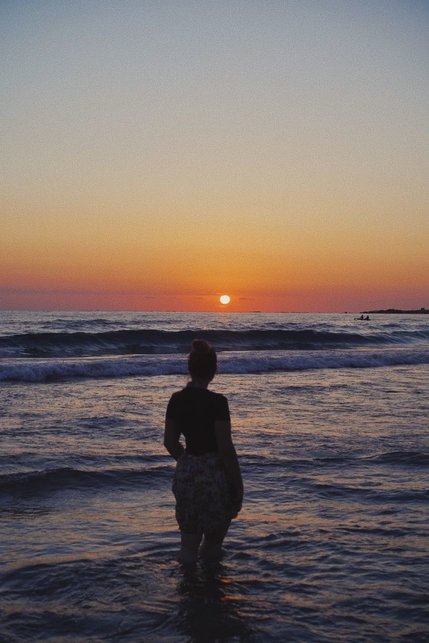 REAR VIEW OF WOMAN STANDING AT BEACH DURING SUNSET