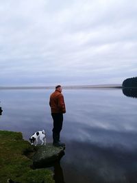 Side view of man with dog standing on rock by lake against sky