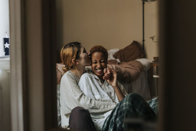 Non-binary person kissing happy friend sitting in bedroom at home