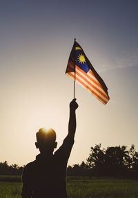 Rear view of man holding flag against sky during sunset