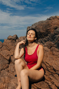 Sensuous young woman with eyes closed sitting on rock against sky