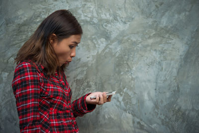 Shocked woman using mobile phone while standing against wall