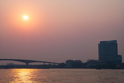 Bridge over river by buildings against sky during sunset