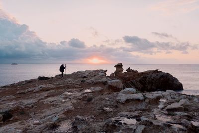 Silhouette man standing on rock by sea against sky during sunset