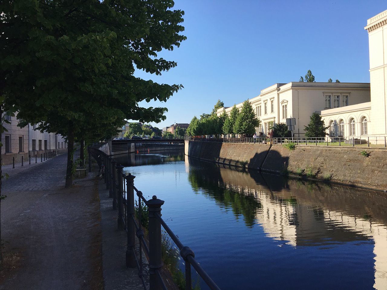 architecture, built structure, building exterior, clear sky, water, tree, railing, blue, sunlight, day, shadow, house, plant, outdoors, sky, canal, incidental people, footpath, copy space, no people