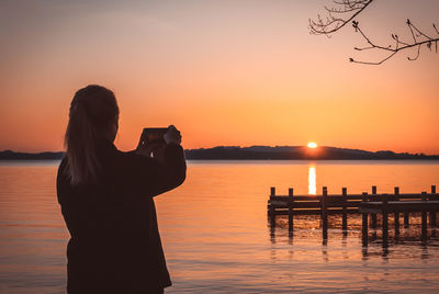 Rear view of woman photographing lake against orange sky