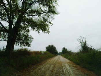 Dirt road amidst trees on field against sky
