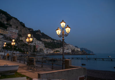 Illuminated street light by building against sky at dusk