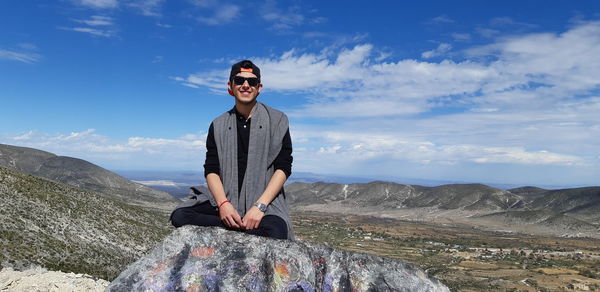 Portrait of smiling man sitting on rock against sky