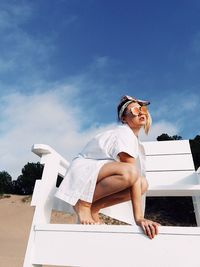 Full length of young woman crouching on lifeguard chair at beach against sky