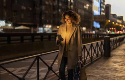 Smiling woman holding shopping bags using smart phone on footpath at dusk