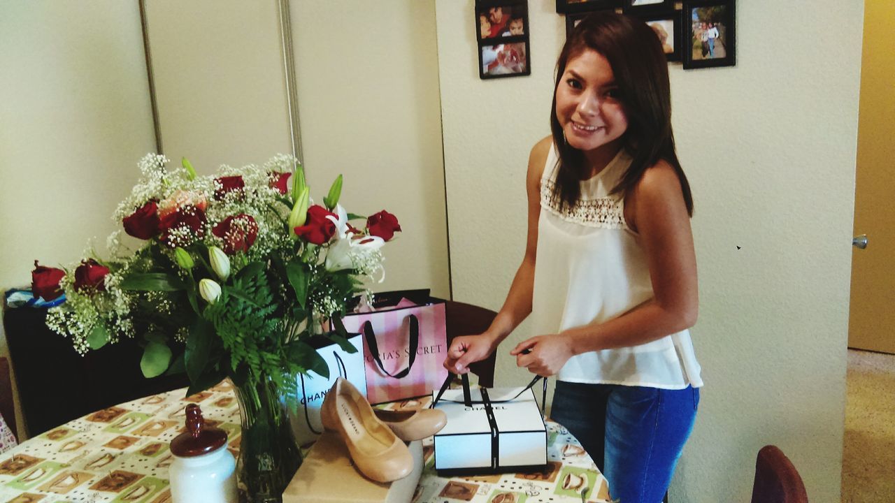 indoors, young adult, casual clothing, lifestyles, person, young women, standing, leisure activity, sitting, front view, looking at camera, holding, flower, three quarter length, portrait, table, smiling, full length