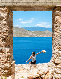 Rear view of woman sitting on rock by sea against sky