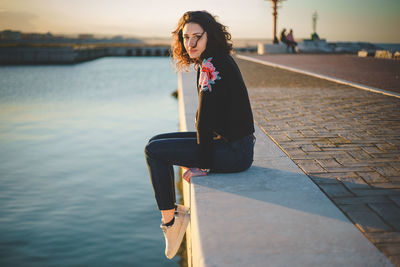 Portrait of woman sitting on retaining wall over sea during sunset