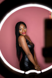 Attractive african american female model in stylish black dress shooting video on smartphone while standing on pink background in studio with glowing ring lamp