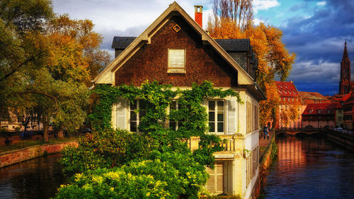 Trees and houses by river against sky during autumn