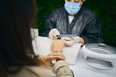 Cropped unrecognizable manicure master using nail file while sitting at table with unrecognizable female client in modern beauty salon during coronavirus pandemic
