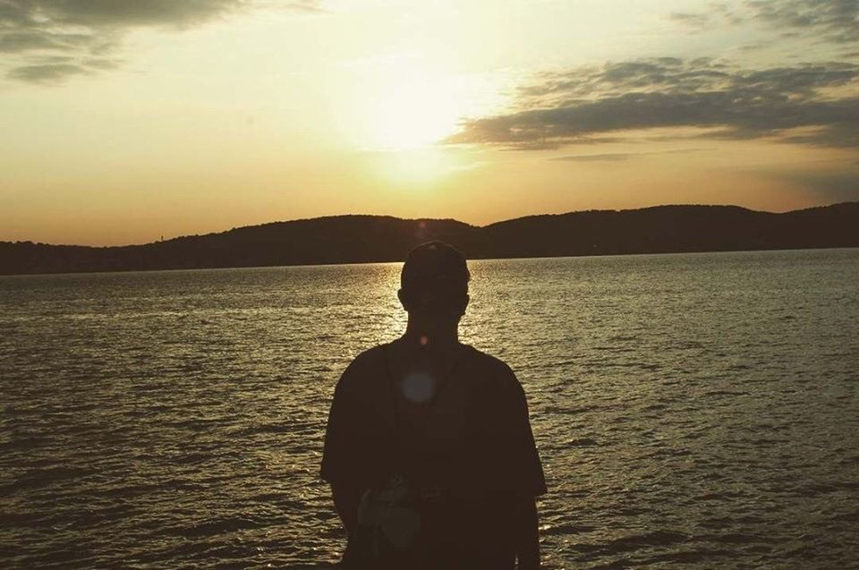 REAR VIEW OF SILHOUETTE MAN STANDING IN SEA AGAINST SUNSET SKY